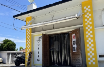 Exterior of a small shop with bright yellow columns featuring white checker patterns, open doorway, and Japanese signage outside. A scooter parked nearby under a clear blue sky.
