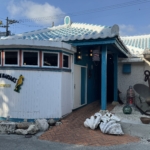 Seafront restaurant with a curved white exterior, turquoise trim, and a blue tiled roof, sea-themed decor on the façade.